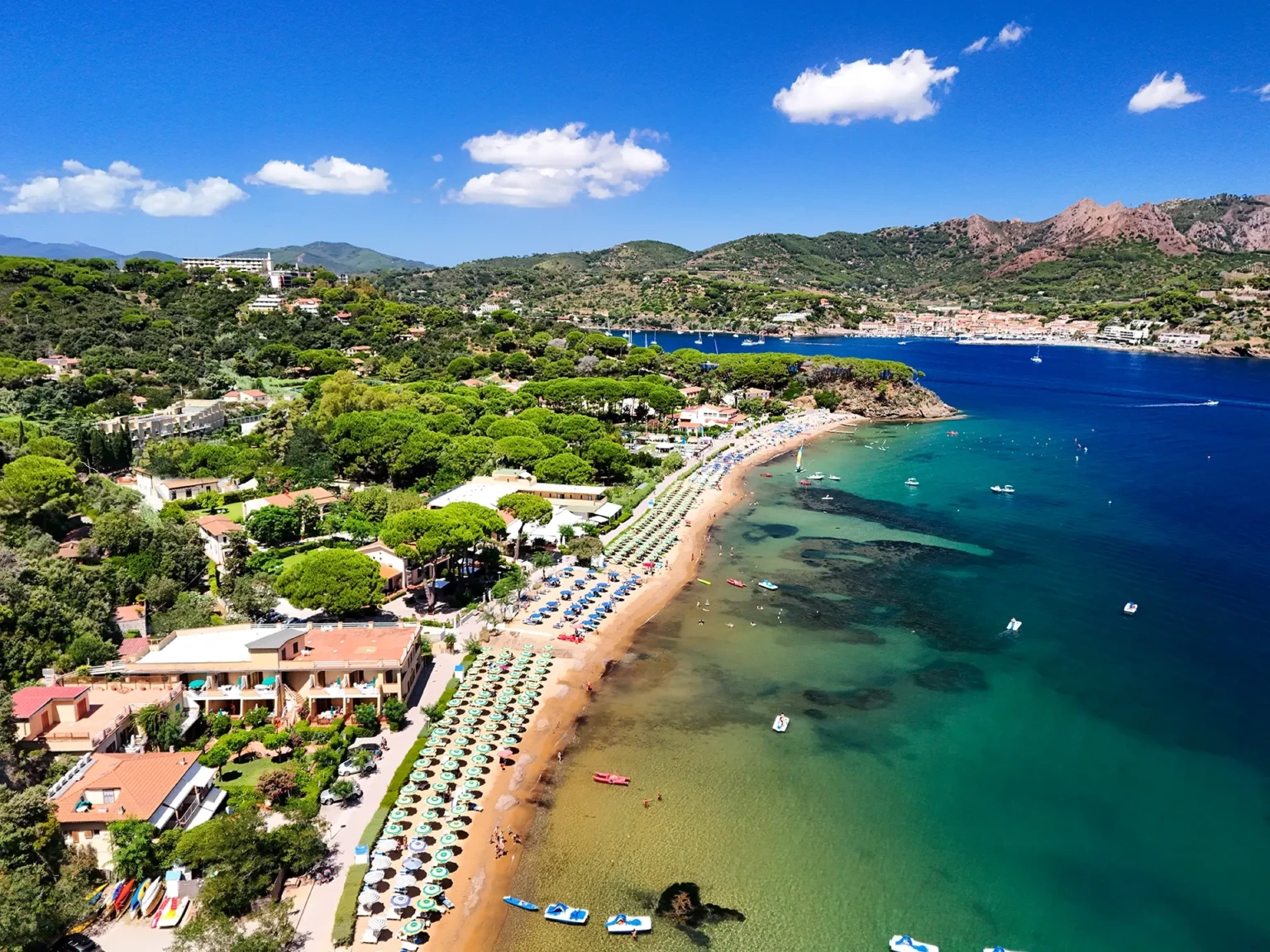 Vista aerea della baia dell'Elba con spiaggia, ombrelloni, hotel e mare turchese tra le colline