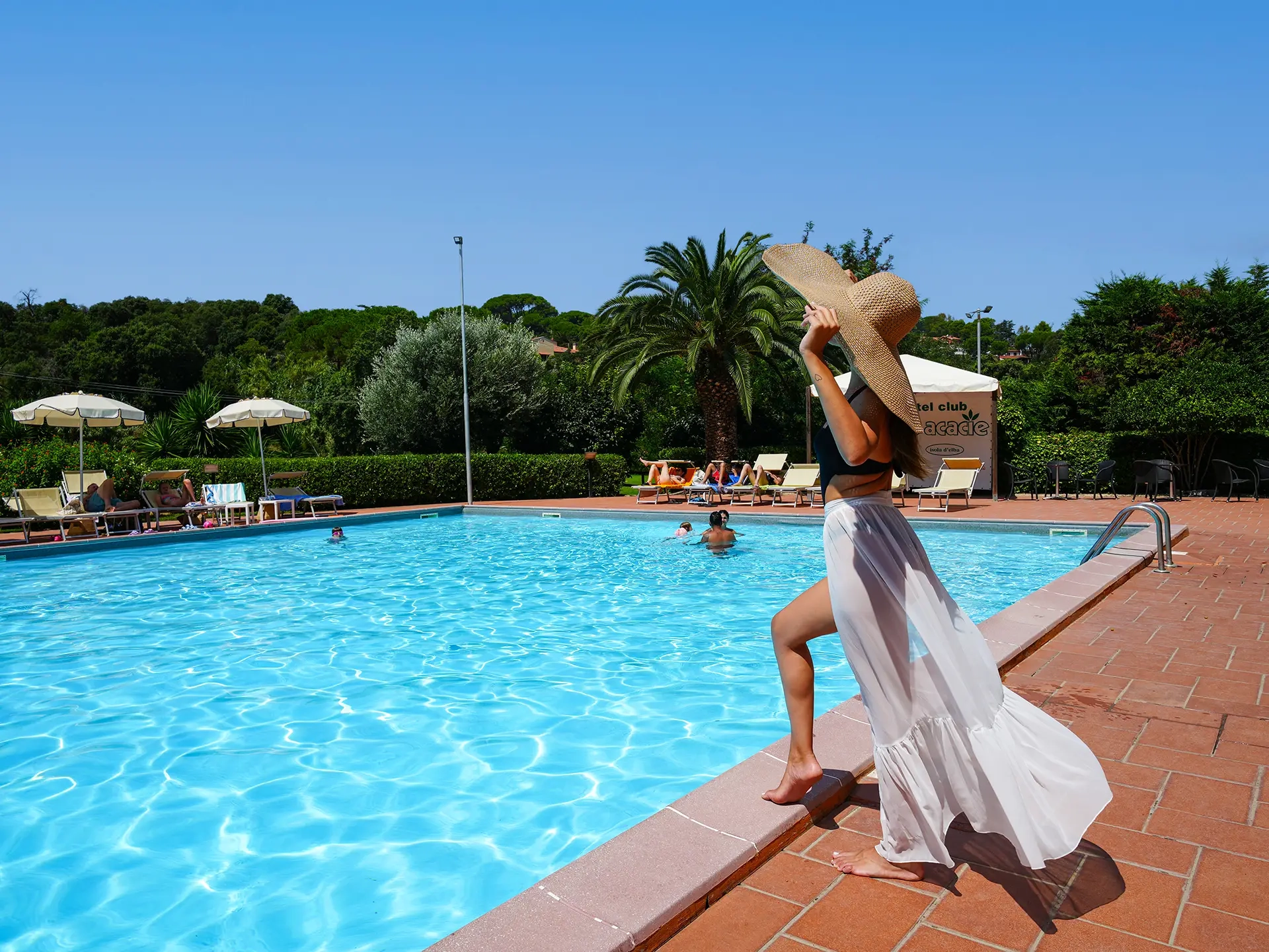 Donna con cappello di paglia e abito bianco a bordo piscina immersa nel verde all'Isola d'Elba