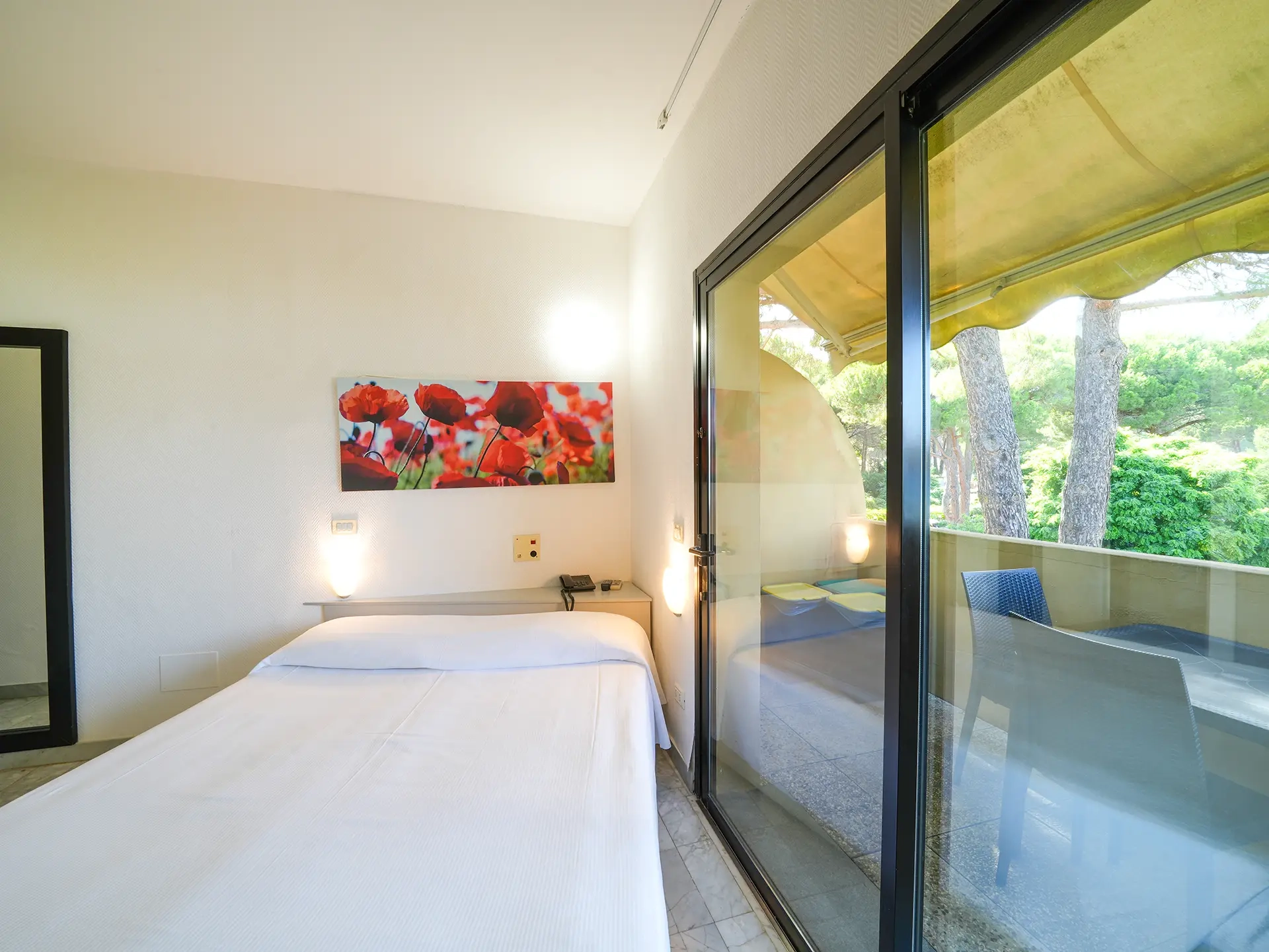 Camera con letto matrimoniale, quadro floreale e ampia porta finestra sulla veranda con vista pineta