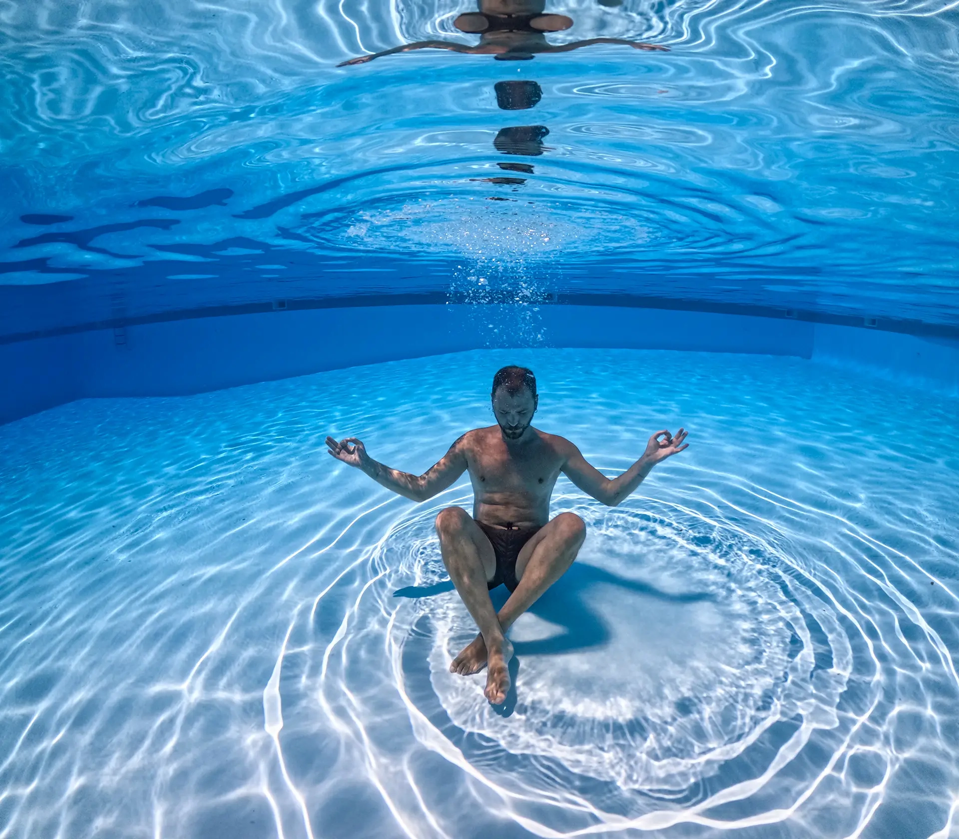 Uomo seduto in posizione yoga sul fondo di una piscina, immerso nell'acqua turchese