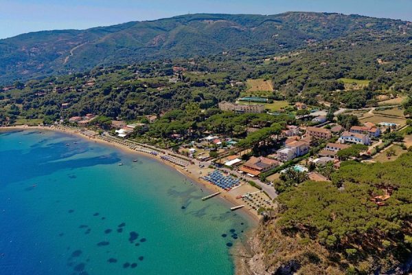 Veduta aerea di una spiaggia e di un villaggio costiero circondato da colline e vegetazione rigogliosa.