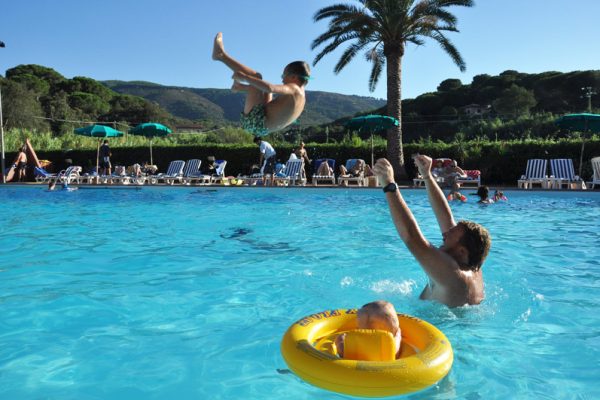 Persone che giocano in piscina sotto il sole, con un uomo che lancia un bambino in aria.