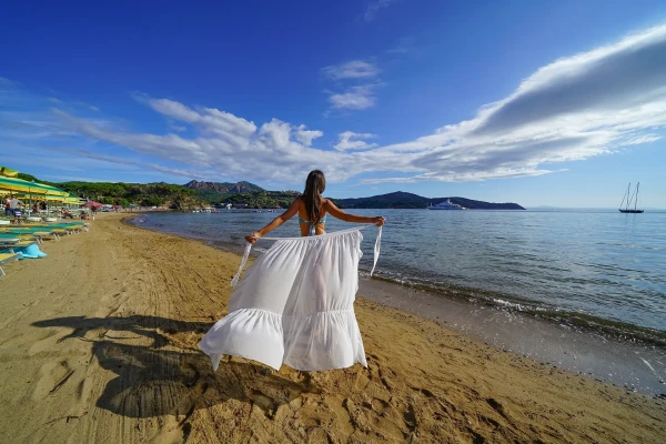 Donna con vestito bianco cammina sulla spiaggia dell'Elba tra ombrelloni e mare turchese